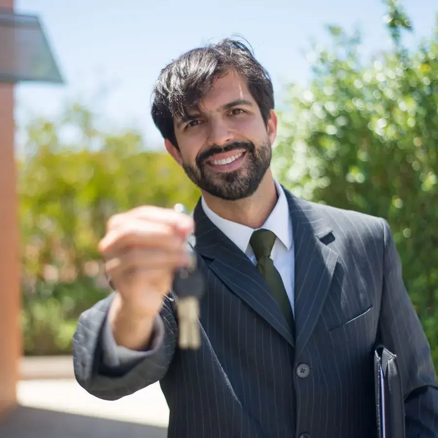 Smiling real estate agent in a pinstripe suit holding out a set of house keys outside a Perth property — Systemly builds automated lead follow-up systems for real estate agents
